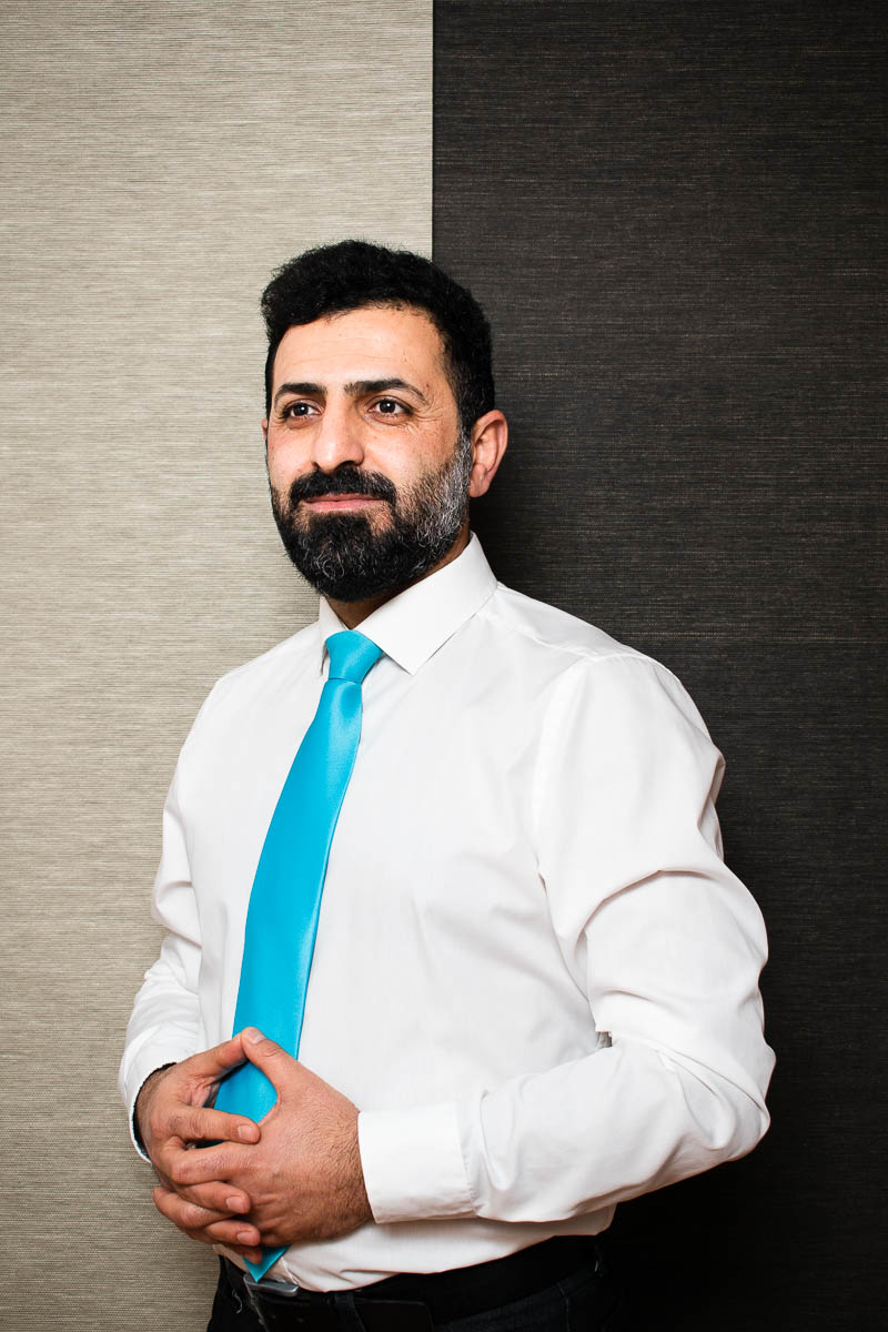 Portrait of refugee Ali in a white shirt and blue tie with his fingers interlocked