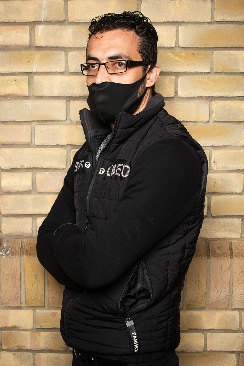 Portrait of refugee Amer standing sideways with his arms crossed wearing a black face mask and jacket