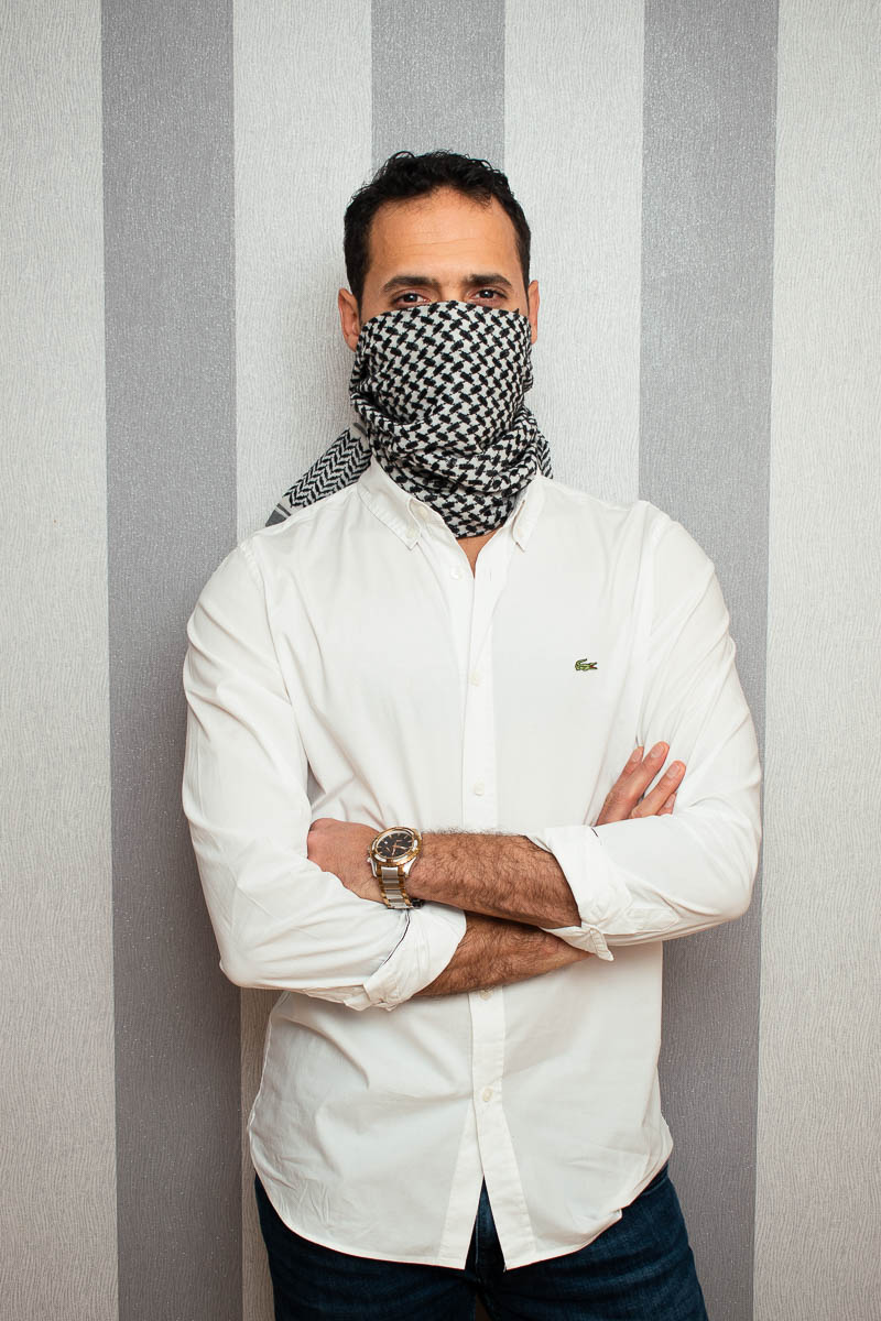 Portrait of refugee Ammar with his hands crossed and lower half of his face covered by a checkered cloth