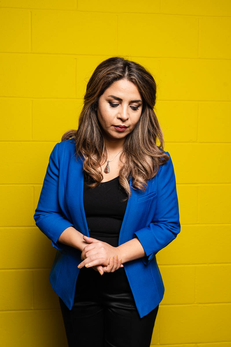 Portrait of refugee Shabnam with her hands clasped and looking down wearing a blue blazer standing against a yellow wall