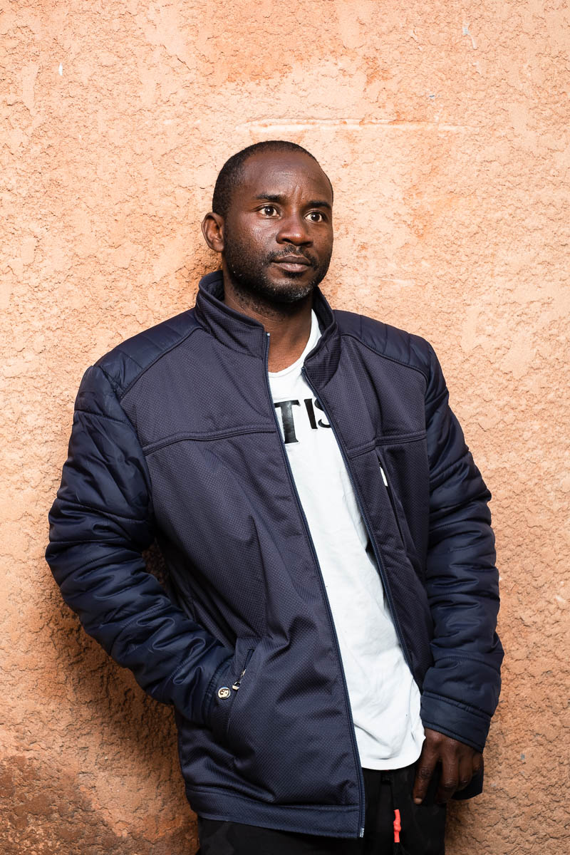 Portrait of refugee Diasson standing with his right hand in the pocket of his black jacket