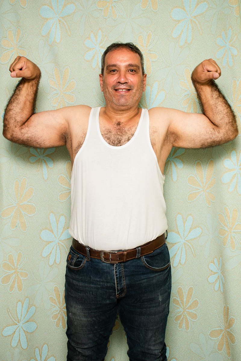 Portrait of refugee Basam wearing a white tank top flexing both his arms by raising them to the sides
