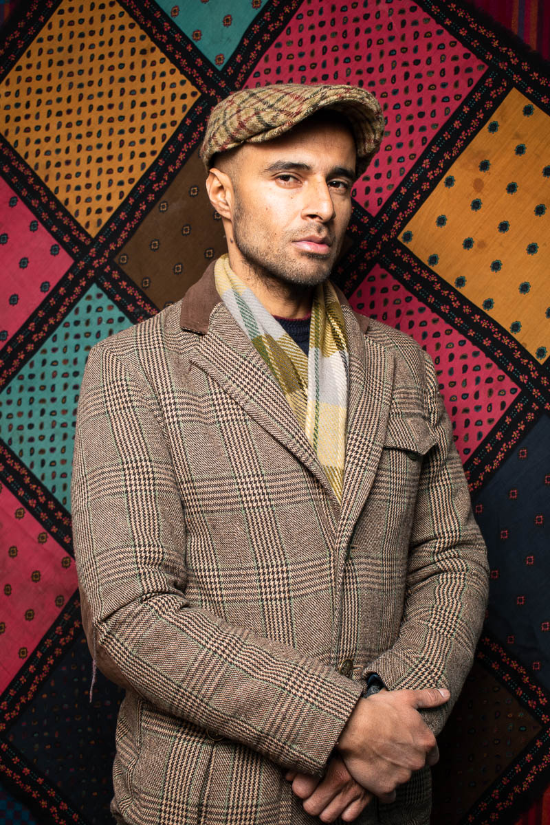 Portrait of refugee Omair wearing a suede coat and a beret, standing with his hands clasped