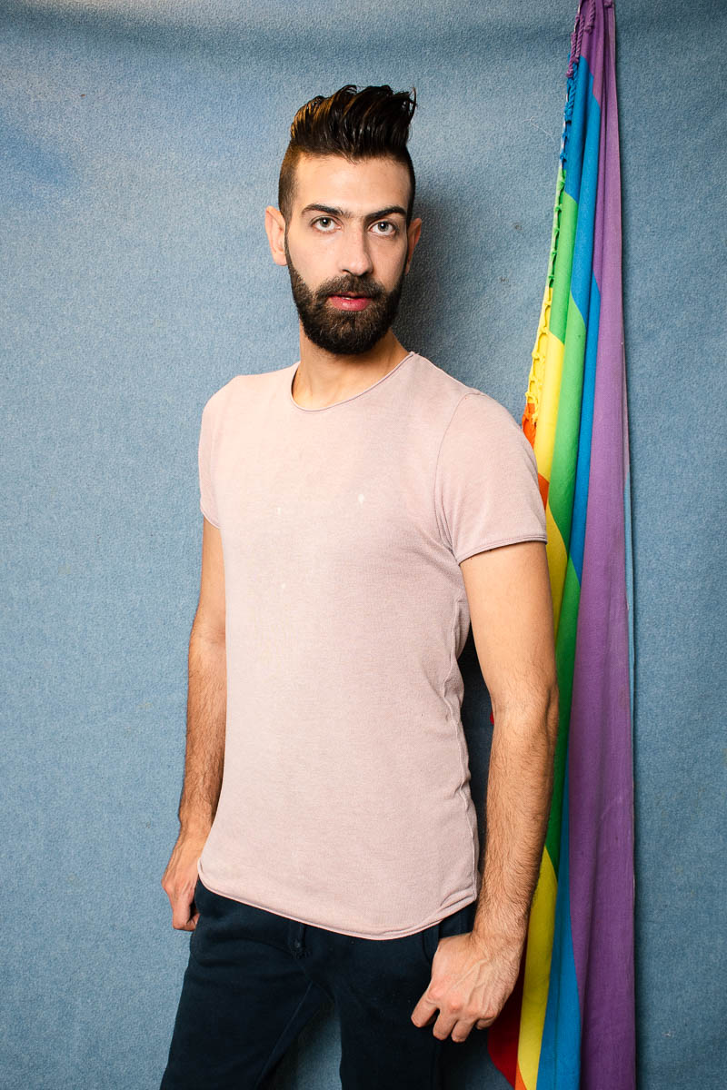 Portrait of refugee Ahmad standing with a rainbow flag mounted behind