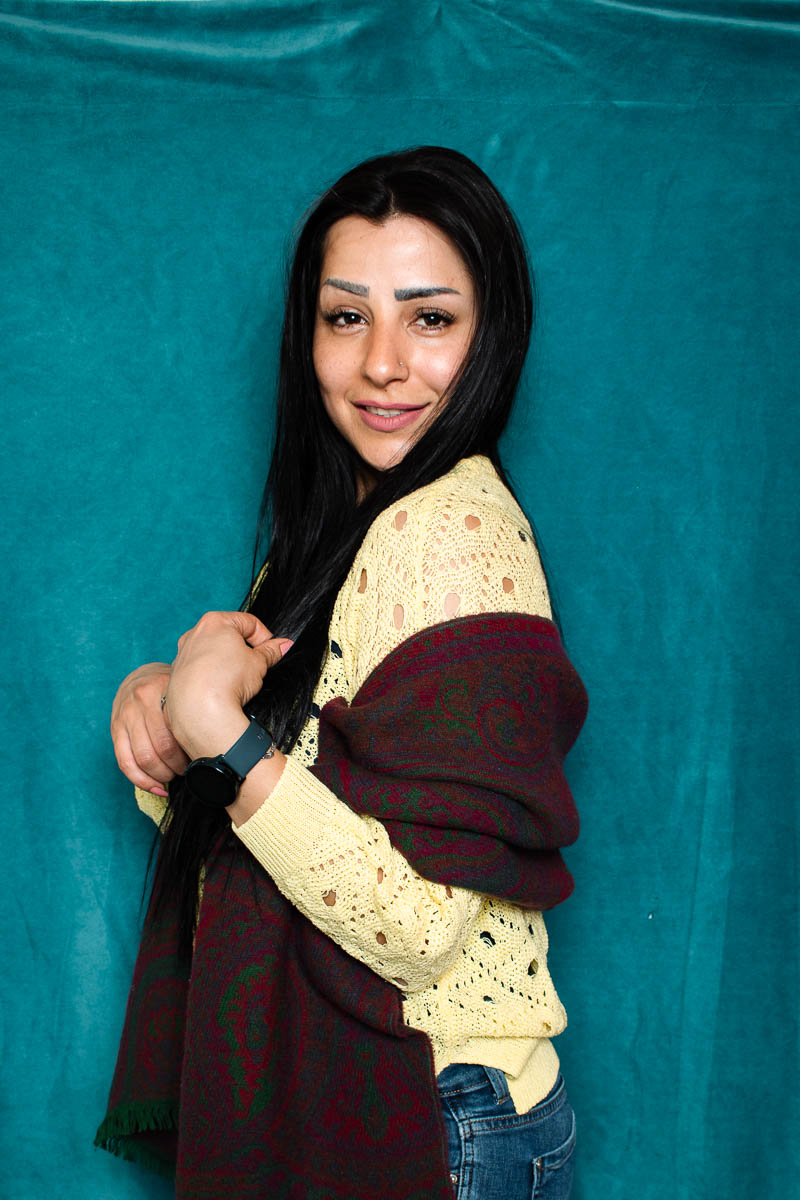 Portrait of refugee Hiam standing sideways using her hands to hold a woollen scarf wrapped around her