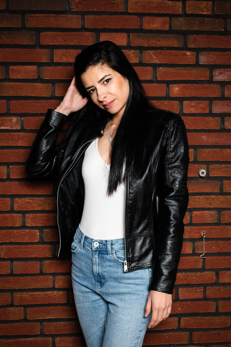 Portrait of refugee Jojo wearing a black leather jacket and one hand holding her hair standing against a brick wall