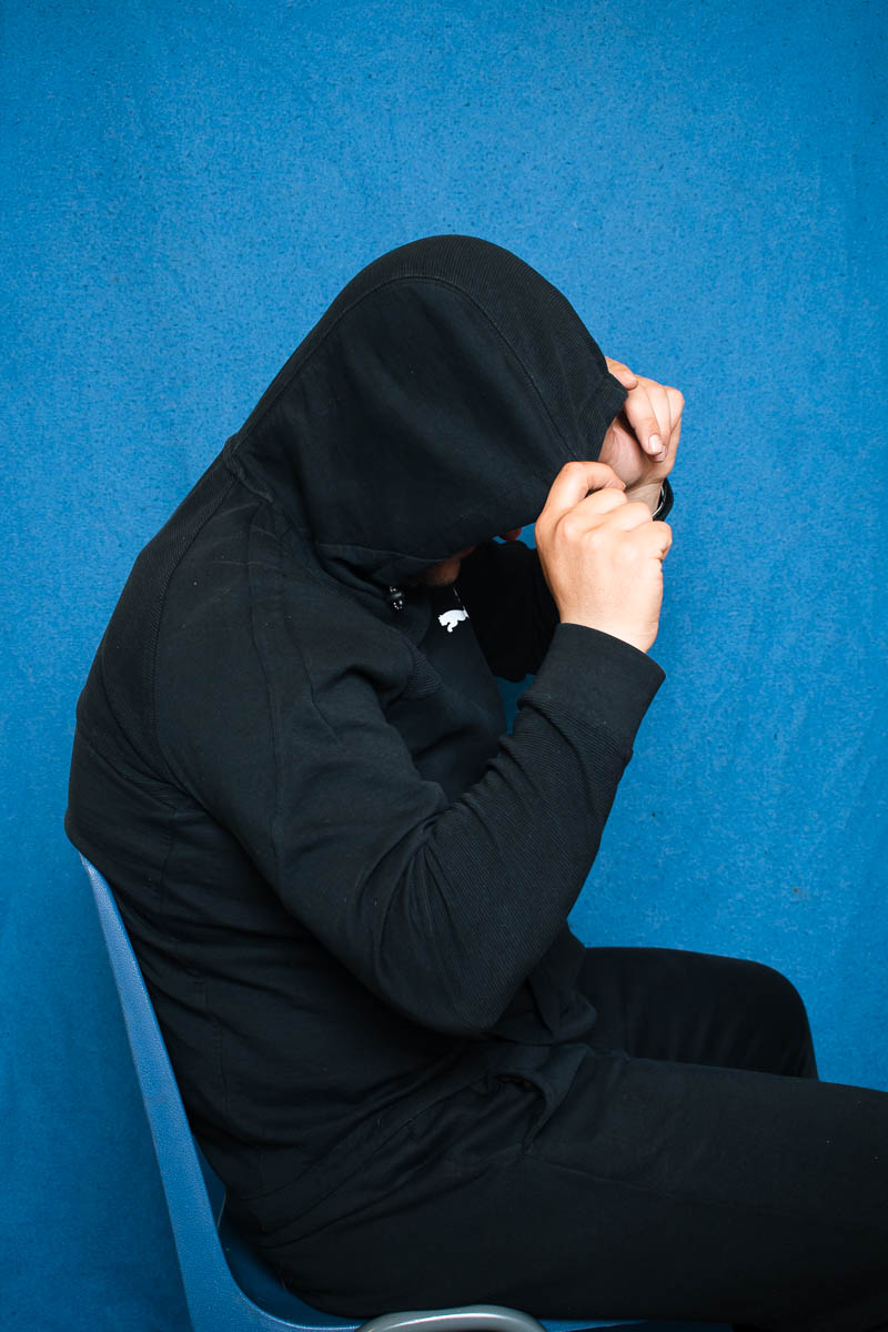 Portrait of refugee Basel sitting on a chair turned to their left hiding their face using the hoodie with both hands