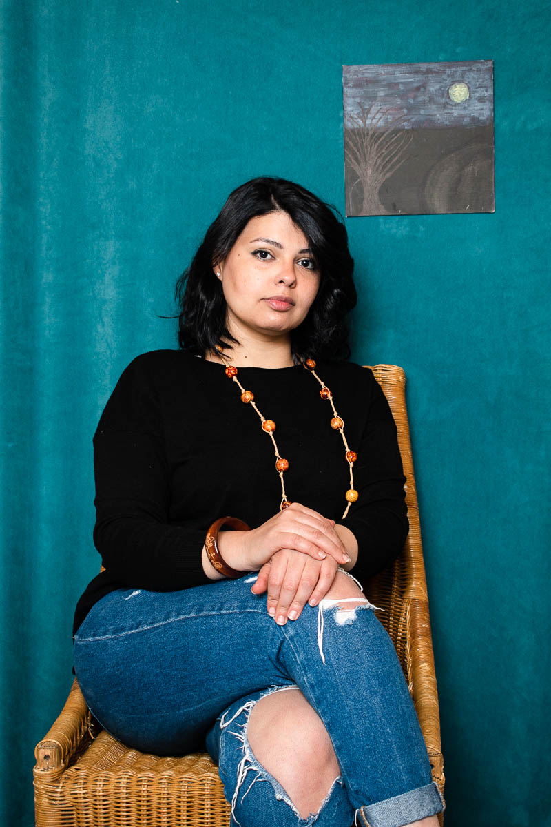 Portrait of refugee Sarah sitting on a straw chair with her hands on top of her folded legs