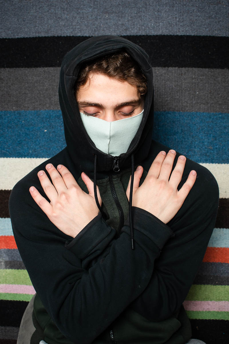 Portrait of refugee Johne looking down with his hands crossed holding both his shoulders wearing a hoodie with the hood up and a face mask