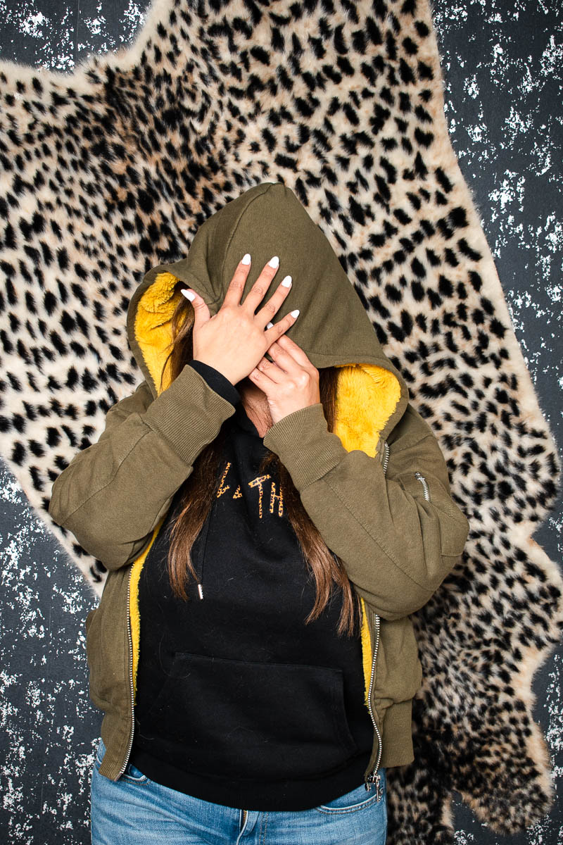 Portrait of refugee Leen with her hands using the hood of her jacket to hide her face standing against a wall with animal printed fur