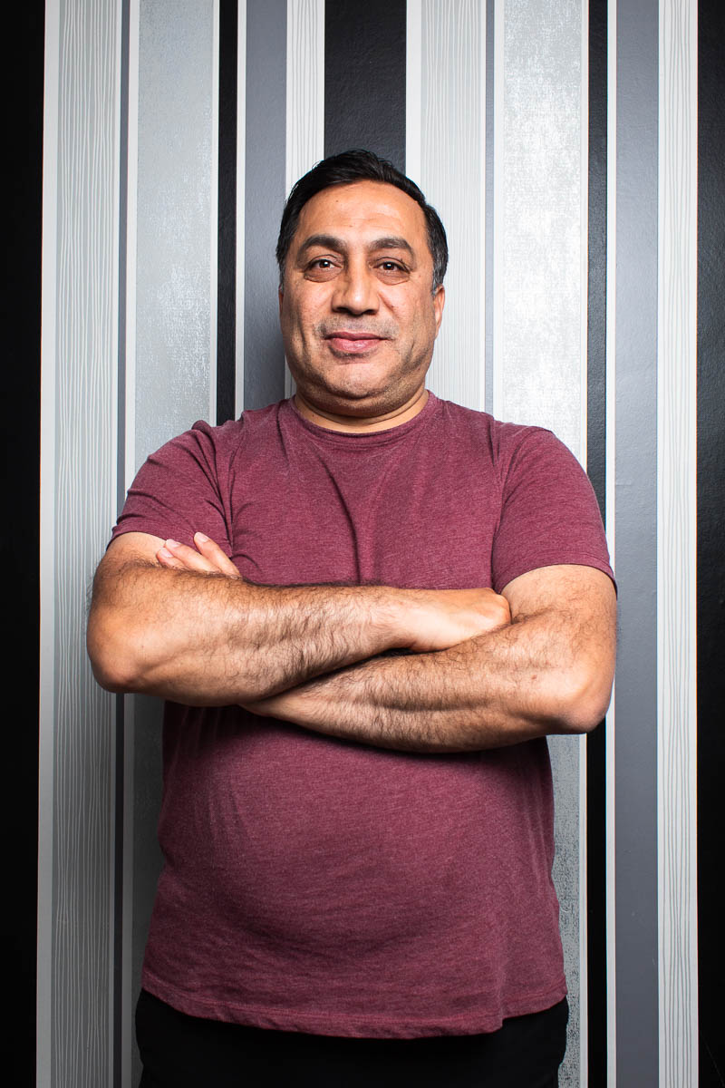 Portrait of refugee Ahmet with his arms crossed wearing a maroon shirt standing against a black and white striped wall