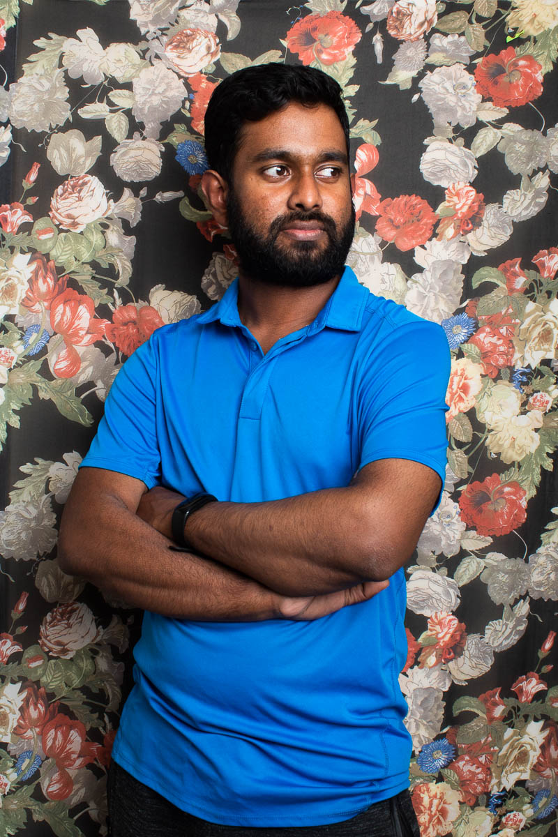 Portrait of refugee Shamil with his arms crossed looking to his left wearing a blue polo shirt standing against a floral background