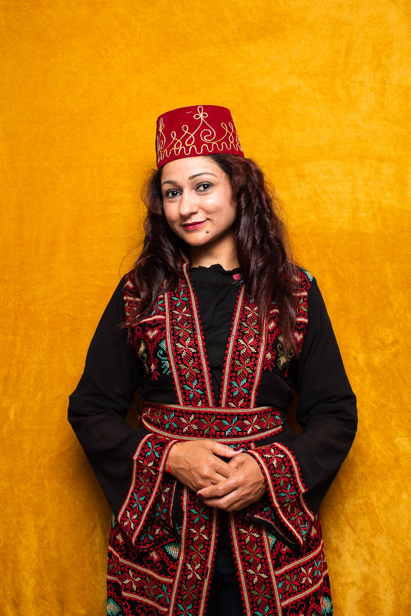 Portrait of refugee Kindah wearing a traditional dress and a fez cap with her hands clasped