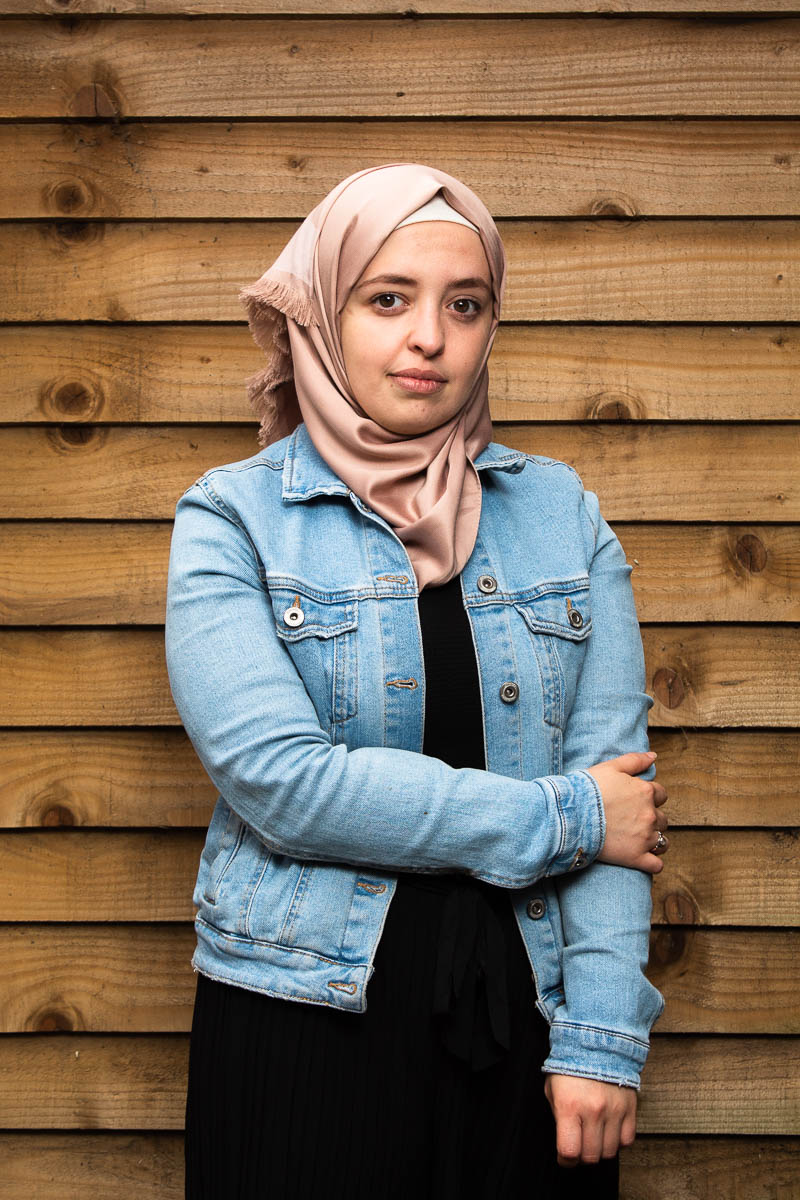 Portrait of refugee Nasma with one hand holding her other arm wearing a denim jacket and hijab standing against a wooden boarded background