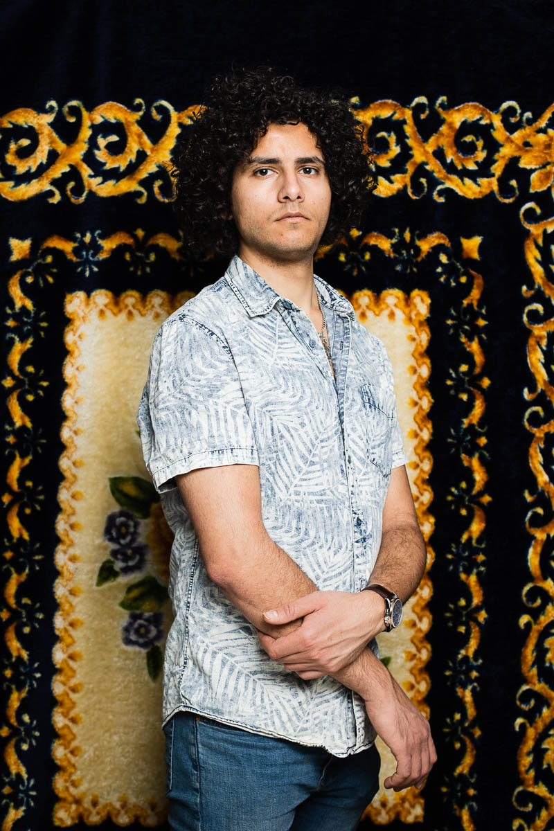 Portrait of refugee Ghazwan with his left hand holding his right standing against a traditionally designed tapestry