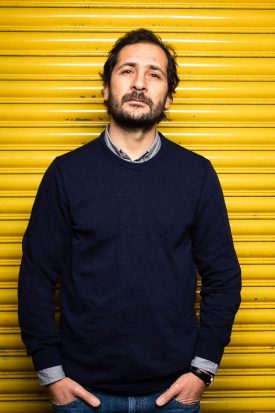 Portrait of refugee Maen with his hands in his pockets standing against a yellow shutter background