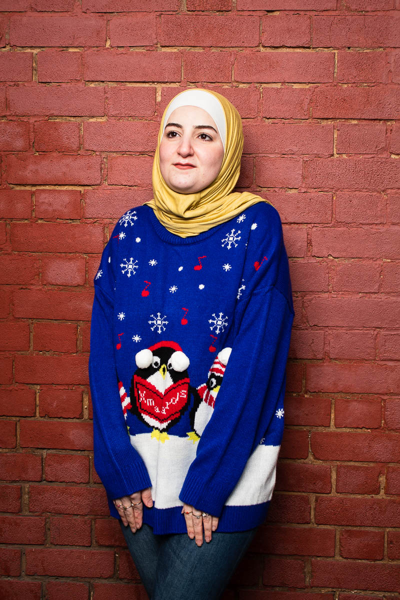 Portrait of refugee Douna wearing a blue Christmas knitted sweater and a yellow hijab standing against a brick wall
