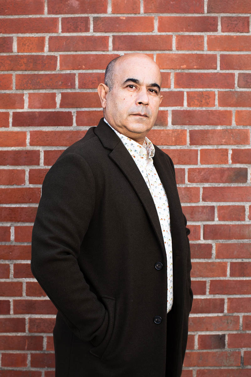 Portrait of refugee Reza standing sideways against a brick wall with his hands in the pockets of his black jacket