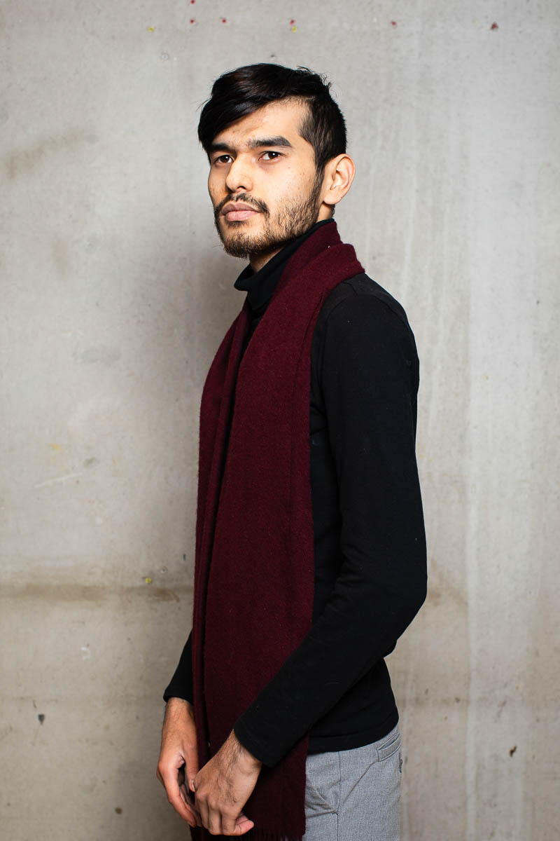 Portrait of refugee standing sideways facing his right with a maroon scarf around his neck