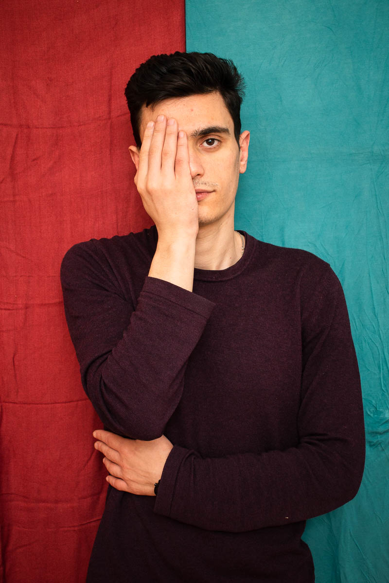 Portrait of refugee Abdo with his right hand covering half his face and the other wrapped around his torso standing against a split red and blue background