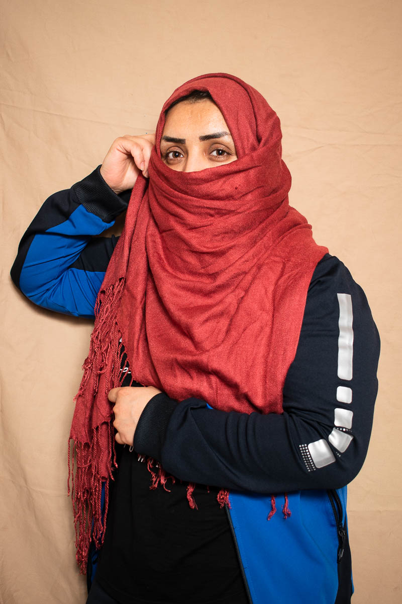 Portrait of refugee Noura with her right hand using her red hijab to cover the lower half of her face