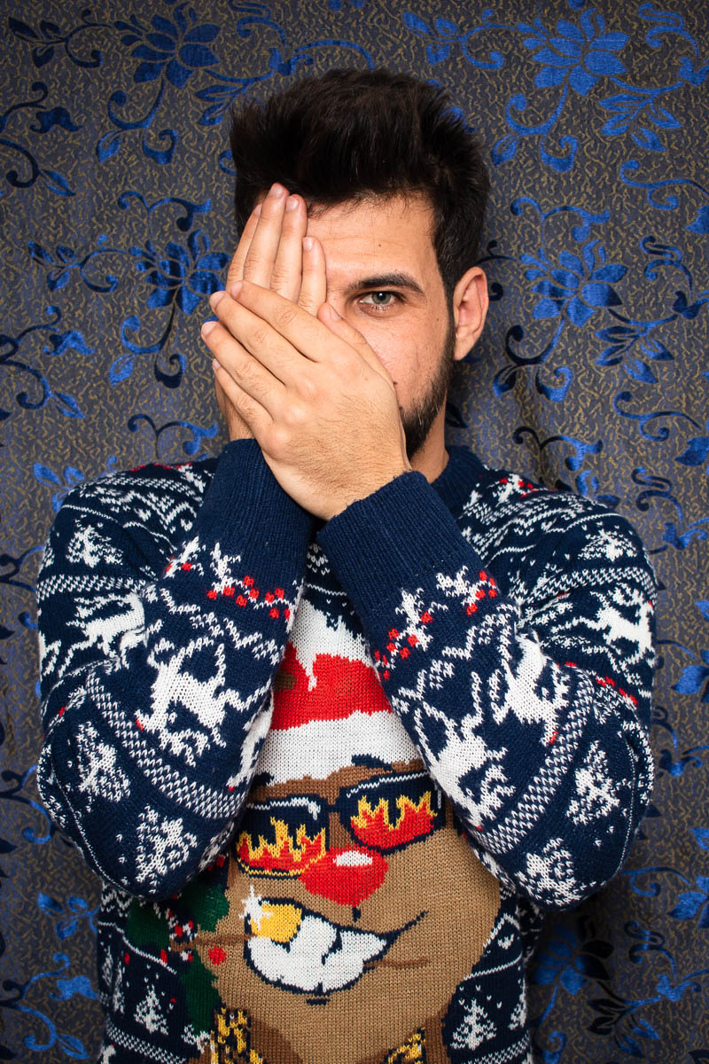 Portrait of refugee Ali hiding his face with his hands except his left eye and wearing a christmas sweater