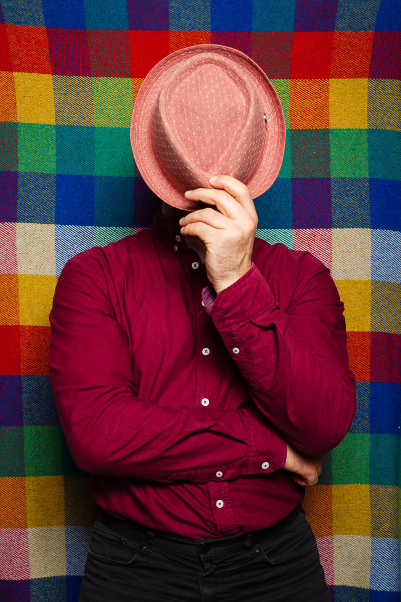 Portrait of refugee Momo using his left hand holding a hat hiding his face standing against a multi-coloured checkered background