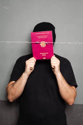 Portrait of refugee Casio hiding his face with a red passport and a barbed wire in front of him