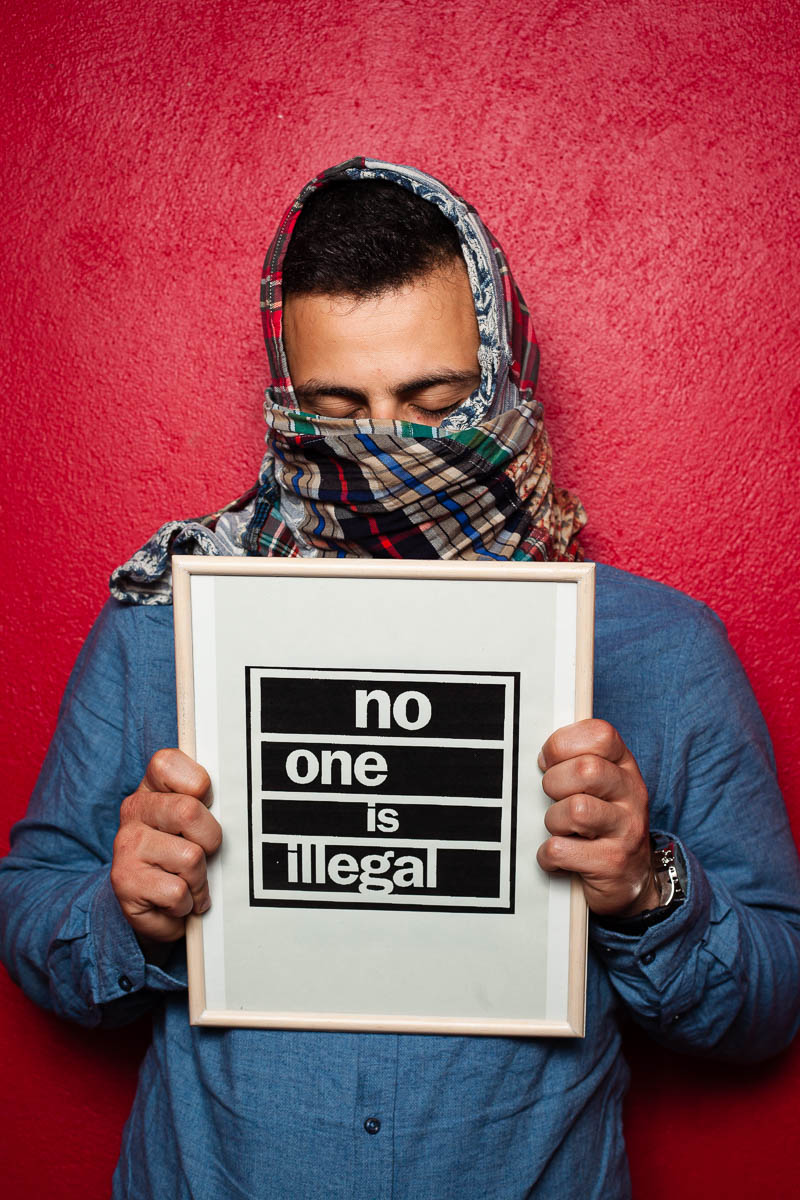 Portrait of refugee Omar with his eyes closed holding up a frame that reads 'no one is illegal' and his face and head covered with a scarf