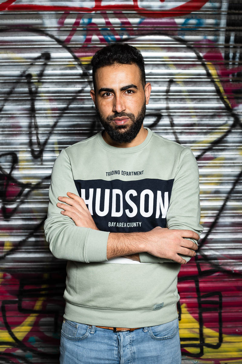 Portrait of refugee Muhammed with his arms crossed standing against a graffiti shutter