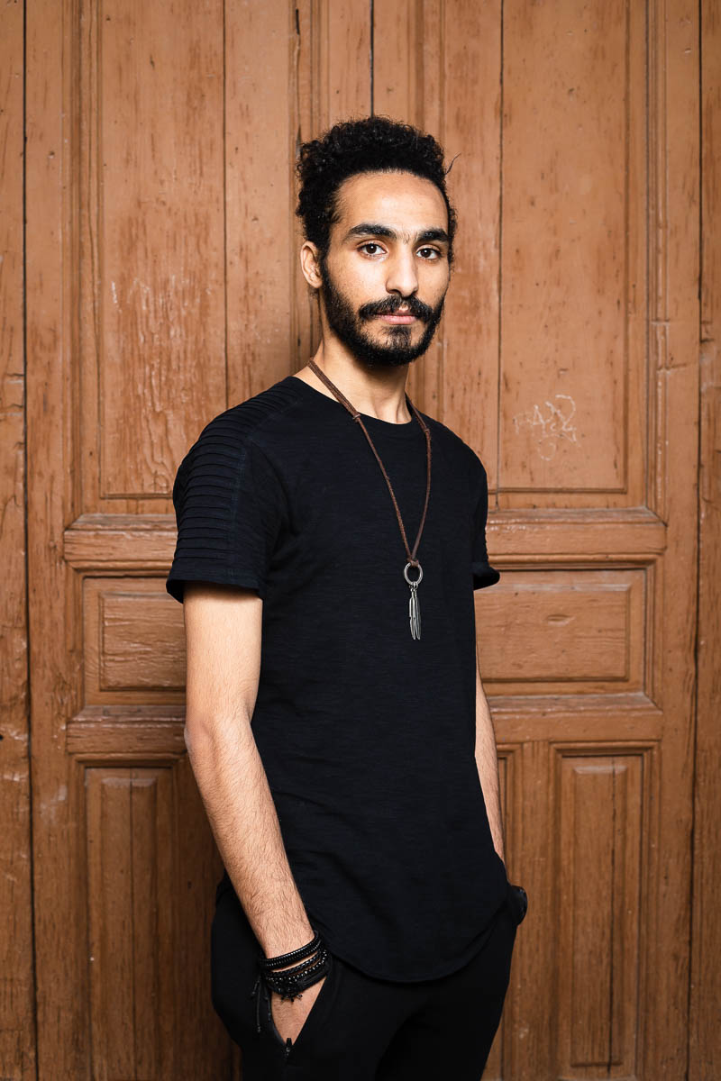 Portrait of refugee Mohamed standing sideways with his hands in his pocket against a wooden door