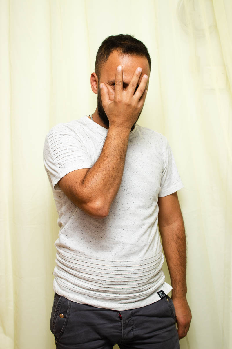 Portrait of refugee Ahmed with his right hand hiding his face and his left thumb in his jean pocket