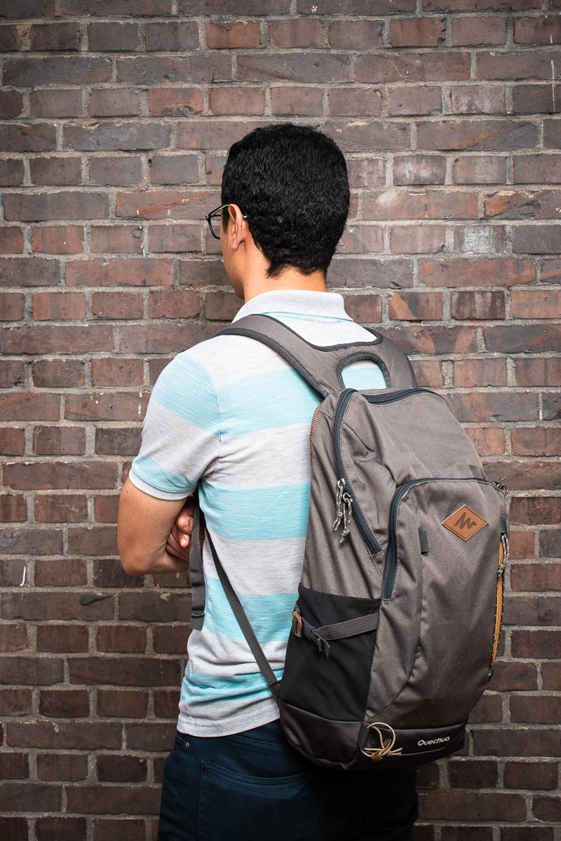 Portrait of refugee Mohamed wearing a backpack with his hands crossed turned away from the camera facing a brick wall
