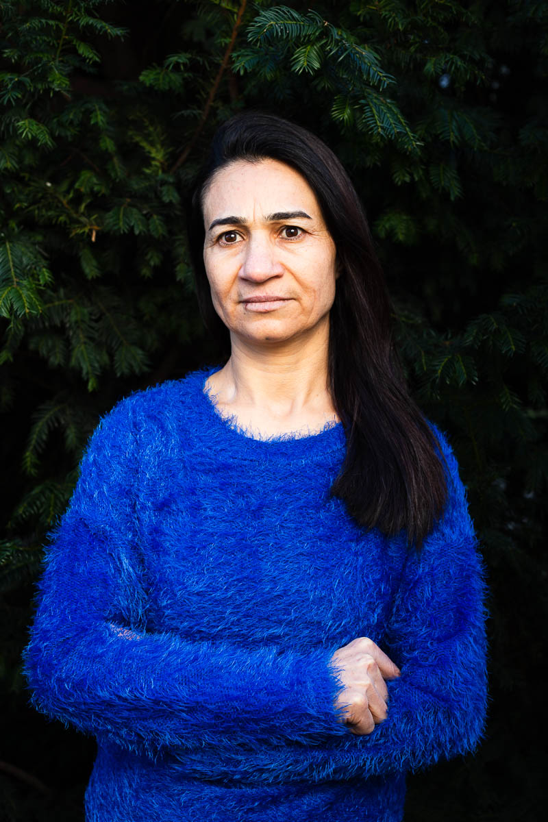 Portrait of refugee Naam wearing a blue woollen sweater with her arms crossed standing against a plant background