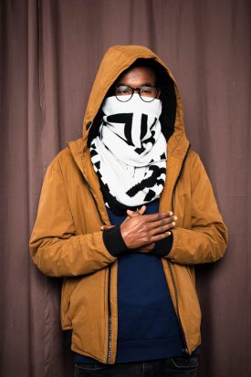 Portrait of refugee Zuhair wearing a brown hoodie jacket and a scarf covering the lower half of his face with his hands crossed and resting on his torso