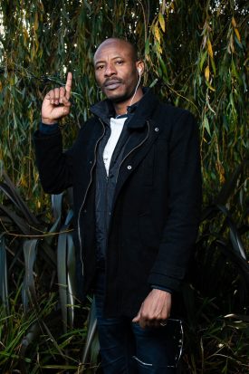 Portrait of refugee Moussa pointing his right forefinger upwards and his left hand holding his sunglasses standing against a tree