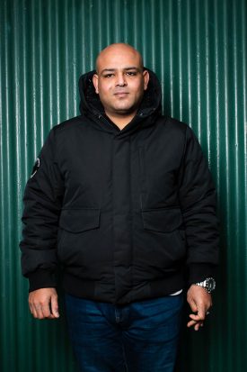 Portrait of refugee Amir wearing a black jacket standing against a dark green shutter