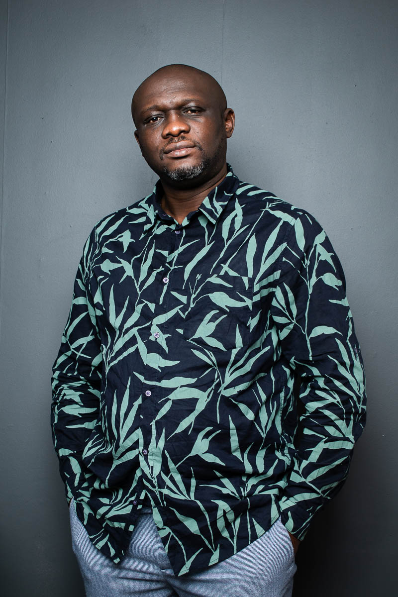 Portrait of refugee Clovis wearing a black patterned shirt with his hands in the pockets of his jeans