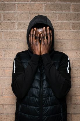 Portrait of refugee Omer wearing a puffer jacket with his hands covering his face standing against a brick wall