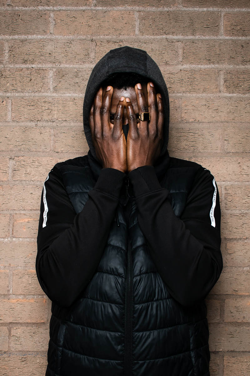 Portrait of refugee Omer wearing a puffer jacket with his hands covering his face standing against a brick wall
