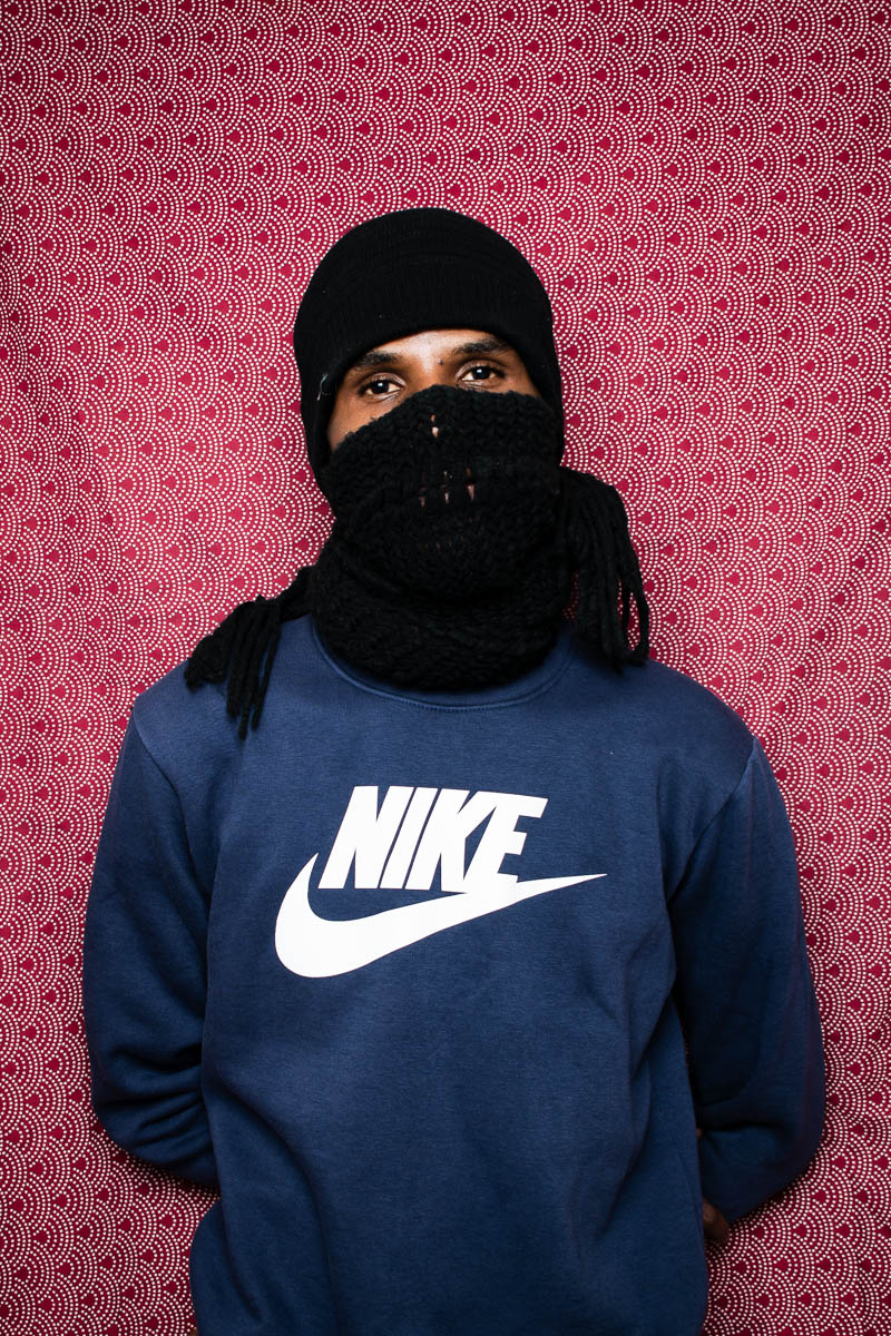 Portrait of refugee Adam with his head and face covered with a black cloth and wearing a blue nike sweatshirt with his hands behind his back.