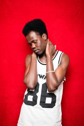 Portrait of refugee Yassin with his hands holding the back of his neck and looking down wearing a basketball jersey