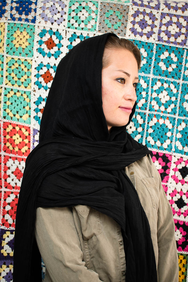Portrait of refugee Zahra turned to her left wearing a black hijab standing against a quilt background