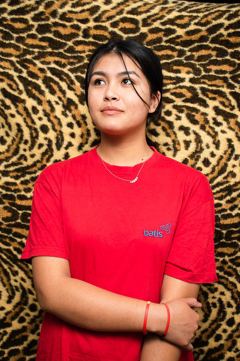 Portrait of refugee Farzaneh with one hand holding her other arm looking to her right standing against an animal printed background