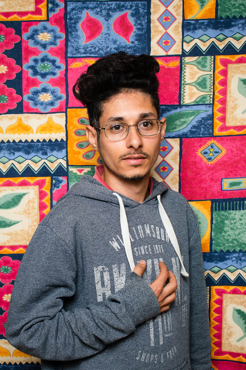 Portrait of refugee Ali wearing a gray hoodie with his right hand on top of his heart standing against a patched up quilt background