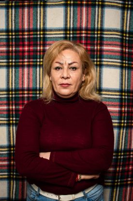 Portrait of refugee Golzar wearing a maroon turtleneck with her arms crossed standing against a plaid background