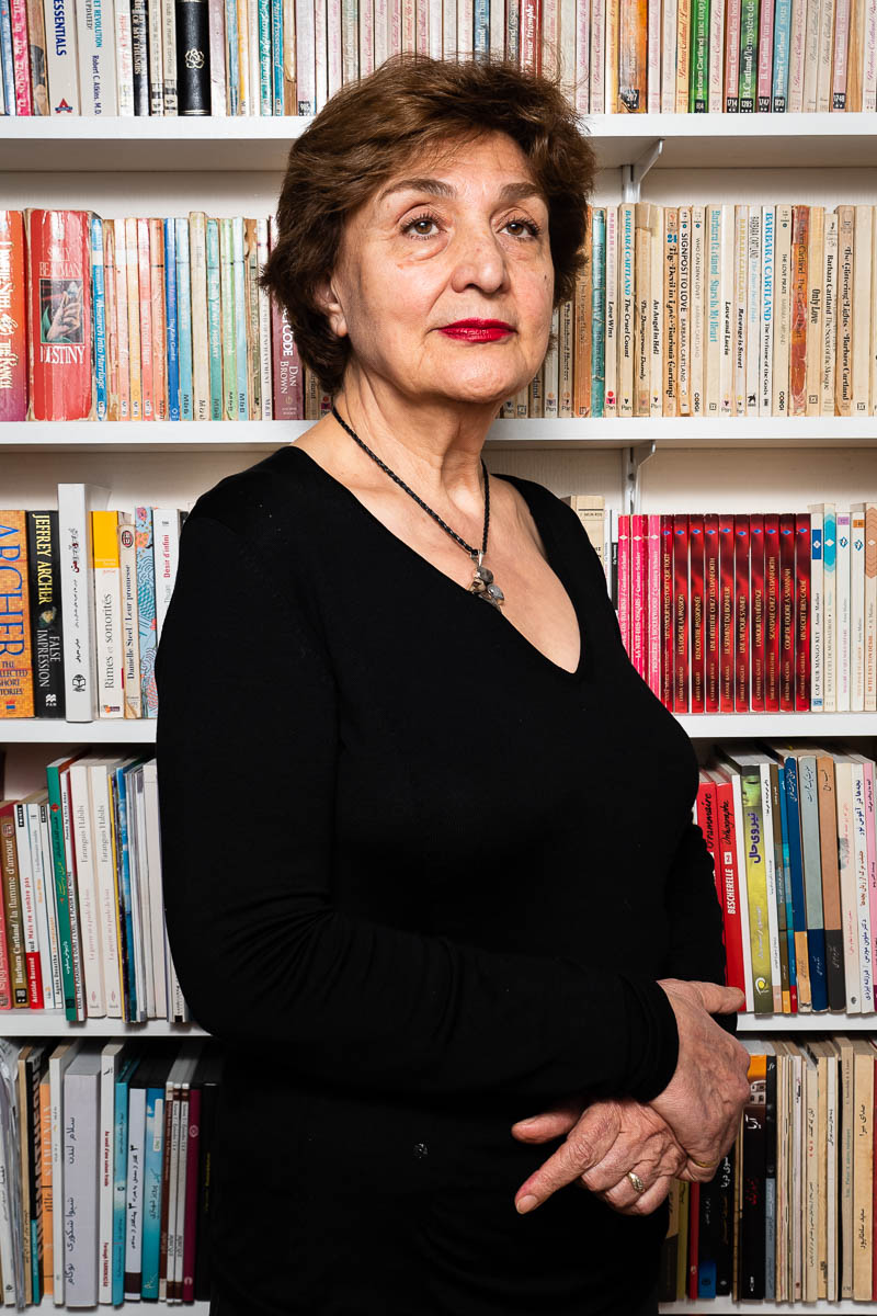 Portrait of refugee Eugénie standing sideways against a bookshelf with her hands folded