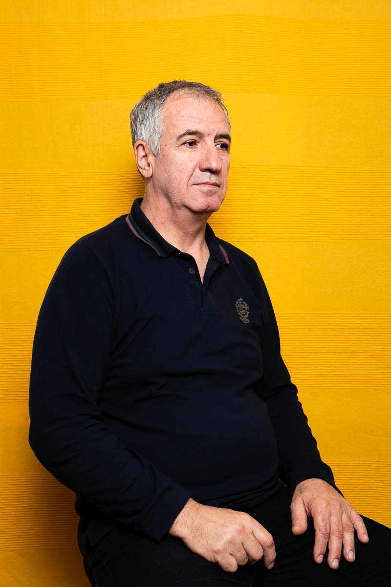 Portrait of refugee Medo sitting with his hands on his lap facing his left against a yellow wall