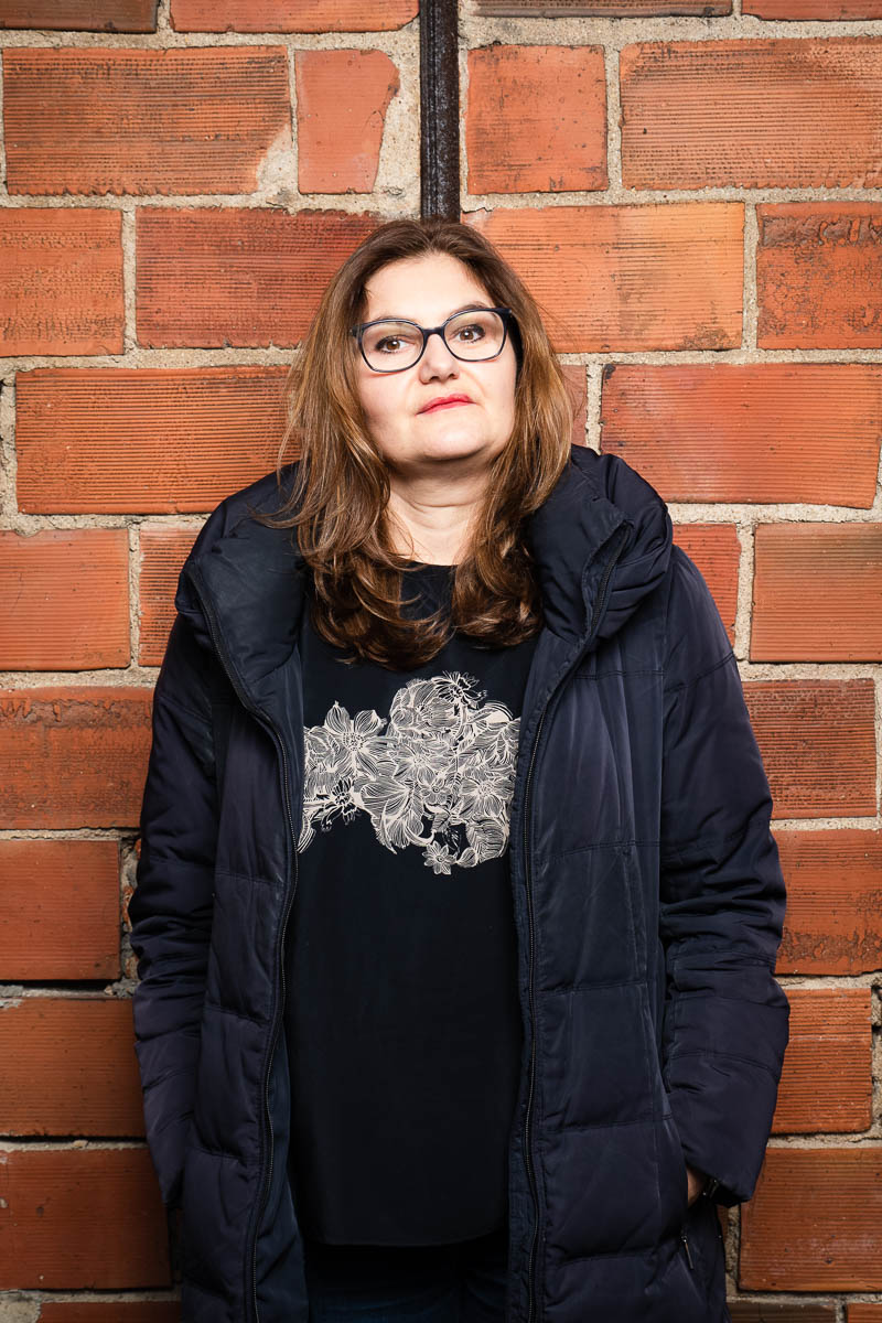 Portrait of refugee Jasna with her hands in the pockets of her black puffer jacket standing against a brick wall
