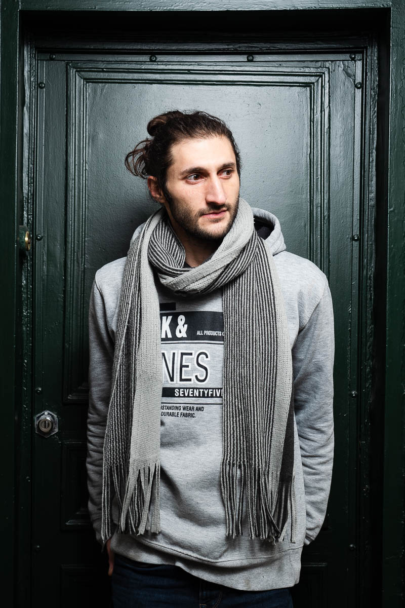 Portrait of refugee Mario in a man bun wearing a thick grey scarf around his neck looking to his left with his hands in his pockets standing against a black door